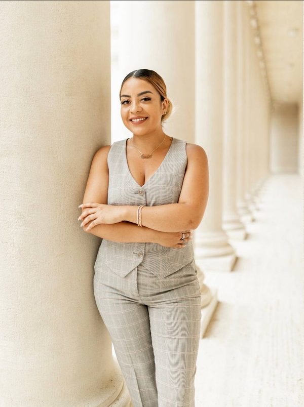 Professional portrait of Jennifer Raya-Gonzalez in a warm, golden-hour lit office. She is wearing a tailored navy blazer and looking directly at the camera with a dignified, reassuring smile. The background features a blurred window view of a city, conveying stability and professionalism.